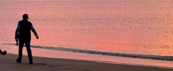 Person walking a dog along a beach shoreline at sunset, with the dog running near the water.